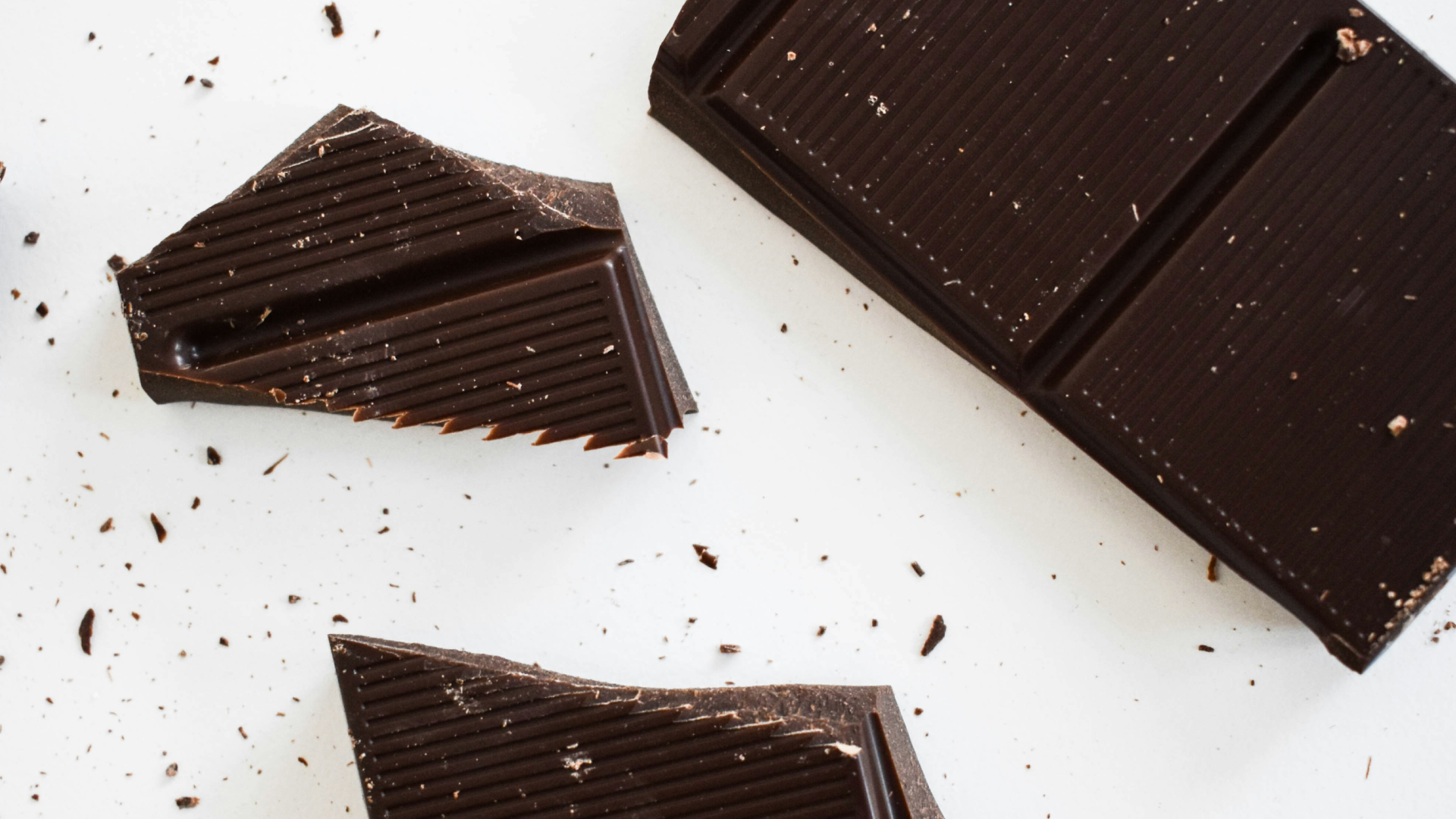 Close-up overhead shot of dark chocolate pieces arranged on a white surface, with broken bar segments and chocolate shavings